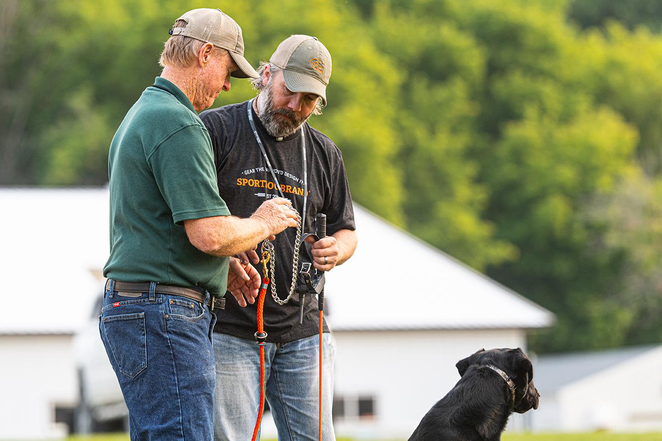 Two men holding SportDOG training gear and a black lab sits next to them 