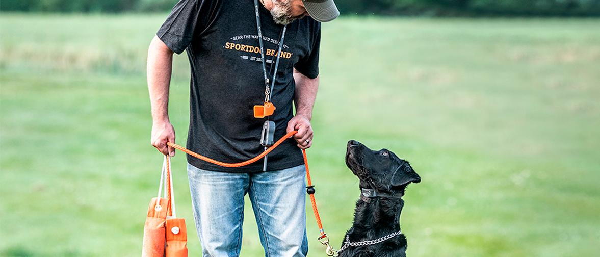 man looking at black lab while holding leash and training dummies. Whistle and e-collar hanging from man's neck.